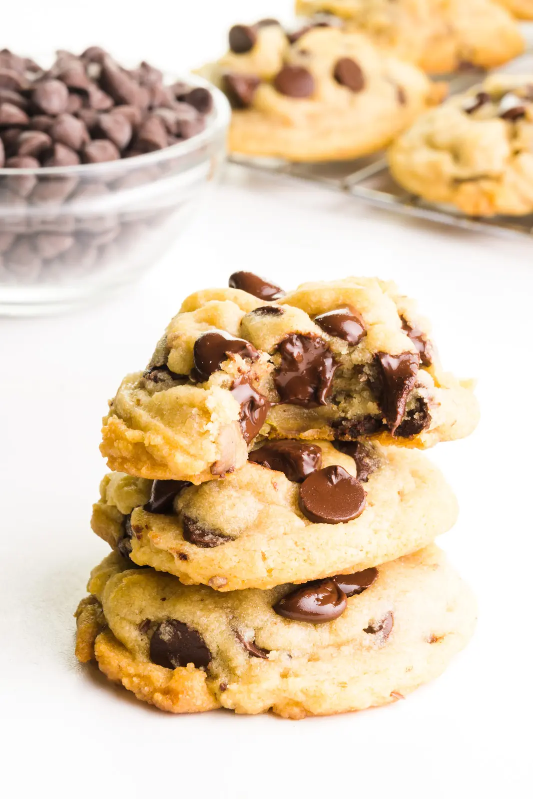 A stack of vegan chocolate chip cookies, shows the top one with a bite taken out. There's a bowl of chocolate chips and more cookies cooling on a wire rack behind it.