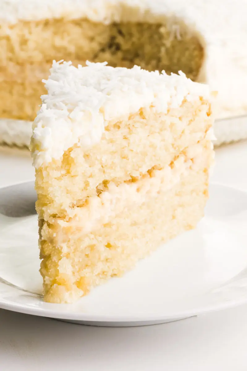 A slice of vegan coconut cake on a plate with the rest of the cake behind it.