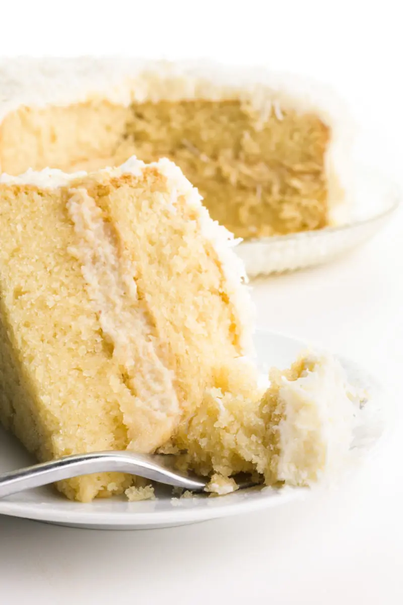 A slice of coconut cake is on a plate with a fork. The fork has a bite of cake on it. The rest of the cake is behind it all.