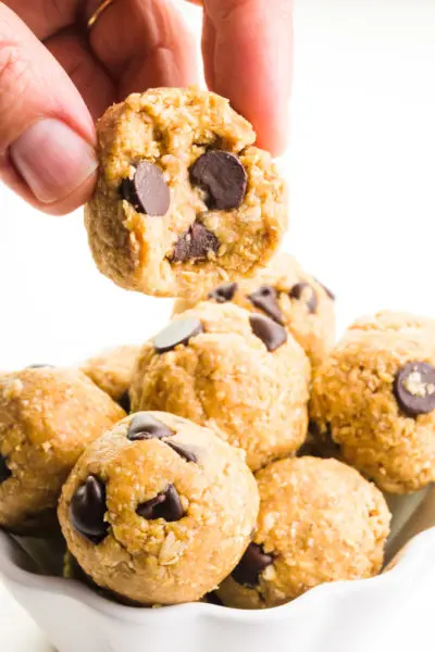 A hand holds a cookie dough ball over a bowl of more of the treats.