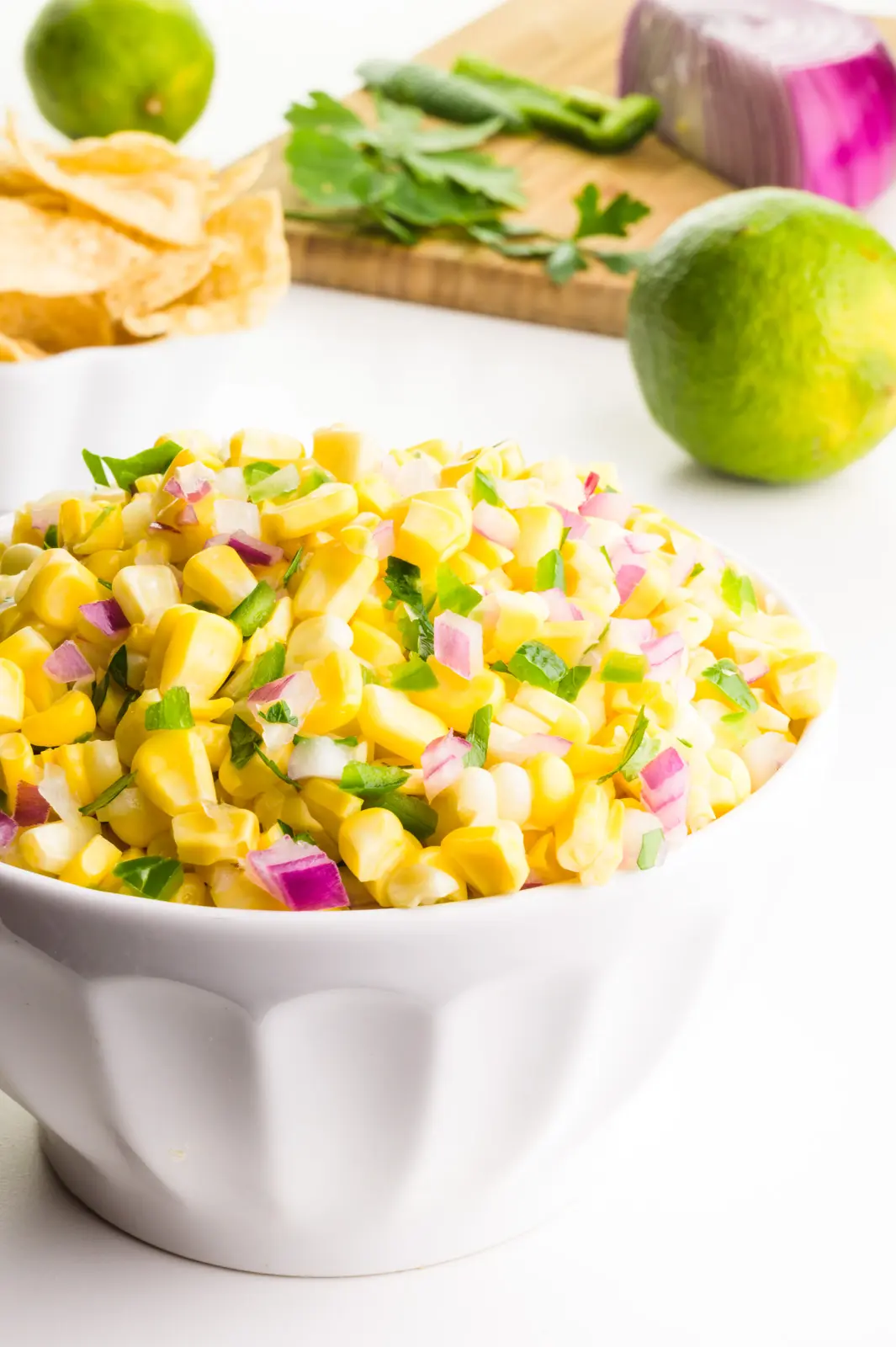 A bowl holds corn salsa. There's a cutting board with ingredients in the background, limes, and a another bowl of tortilla chips.