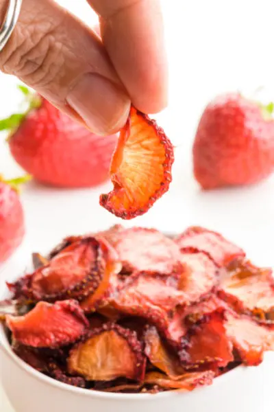 A hand holds a dried strawberry over a bowl of dried strawberry slices. There are fresh strawberries behind the bowl.