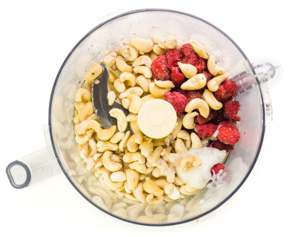 Ingredients such as cashews and raspberries are in the bottom of a food processor bowl.