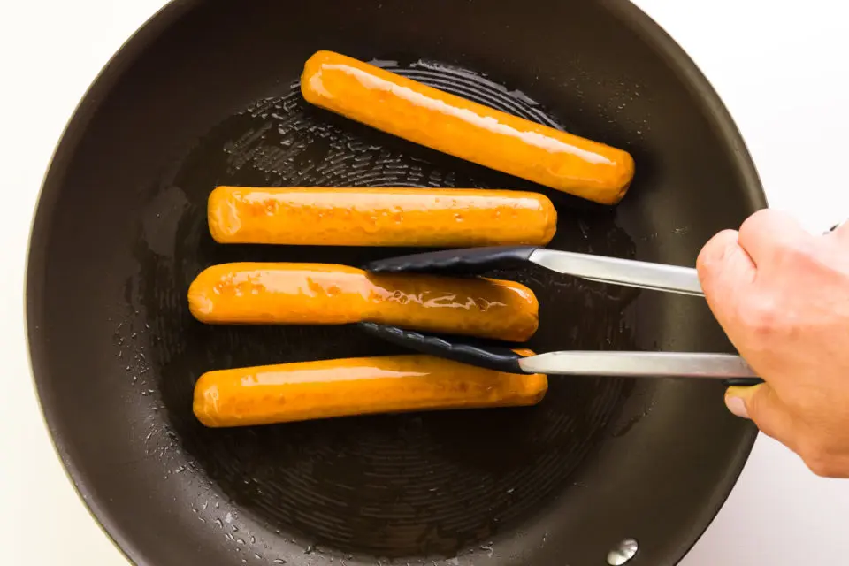 A hand holds tongs, turning vegan hot dogs in a skillet.