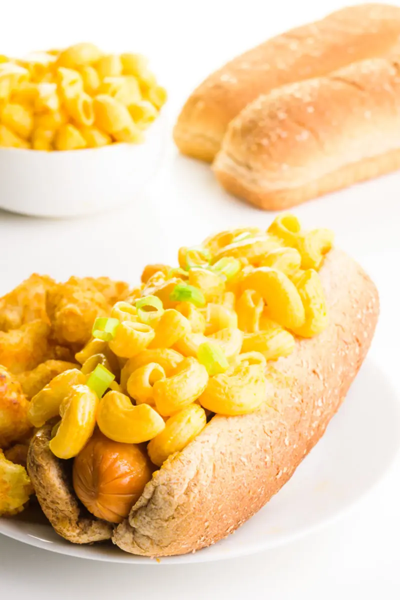 A vegan hot dog has macaroni and cheese on top. There are tater tots next to the hot dog. A bowl of mac and cheese and more hot dog buns are behind it.