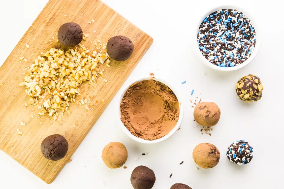 Chocolate truffles sit next to bowls of cocoa powder and sprinkles. A cutting board has chopped nuts on it.