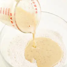 A hand holds a pyrex measuring cup full of a yeast mixture. It's being poured into a flour mixture.