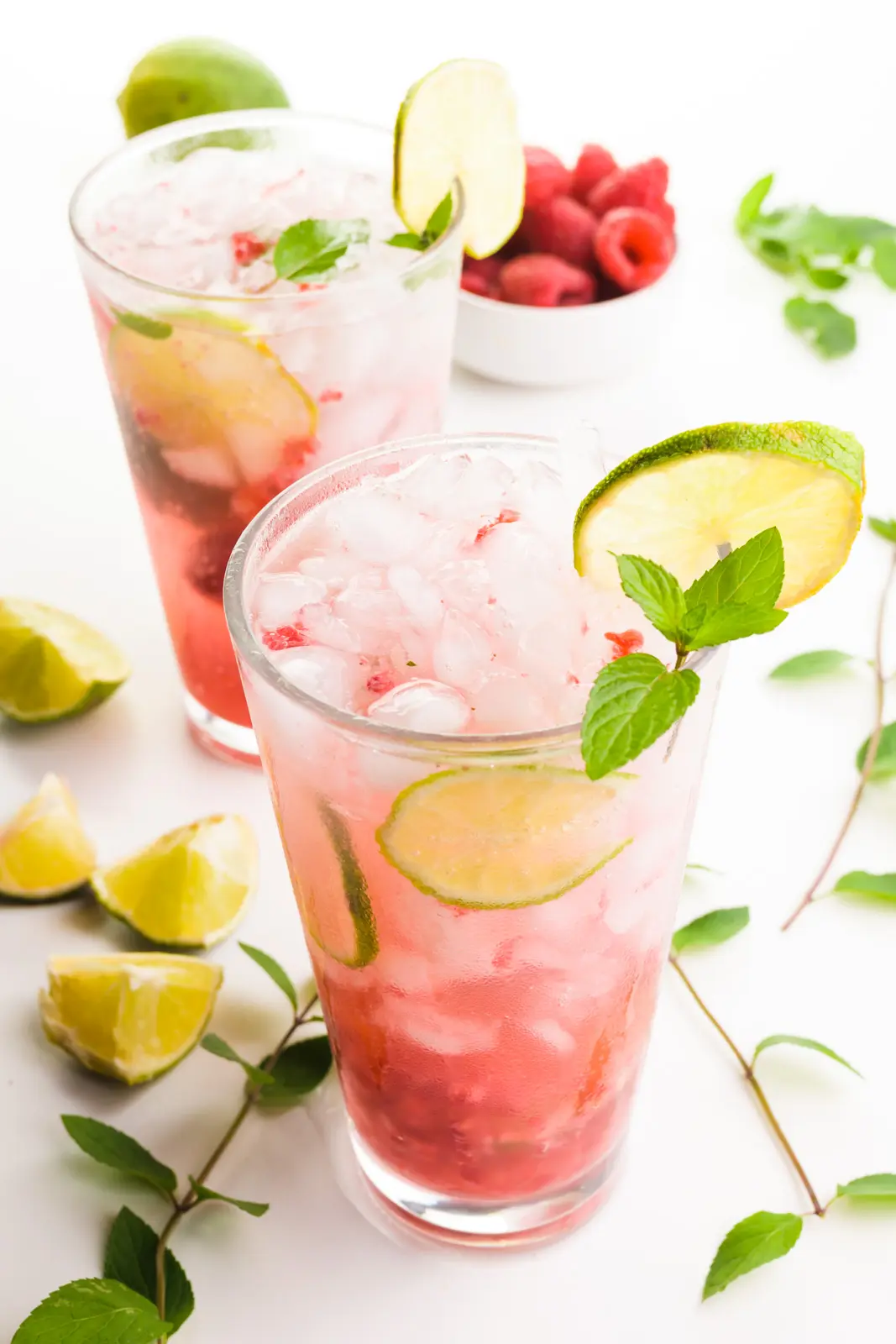 Two tall glasses full of raspberry mojitos have lime wedges as garnishes. There are mint sprigs, limes, and raspberries around the glasses.