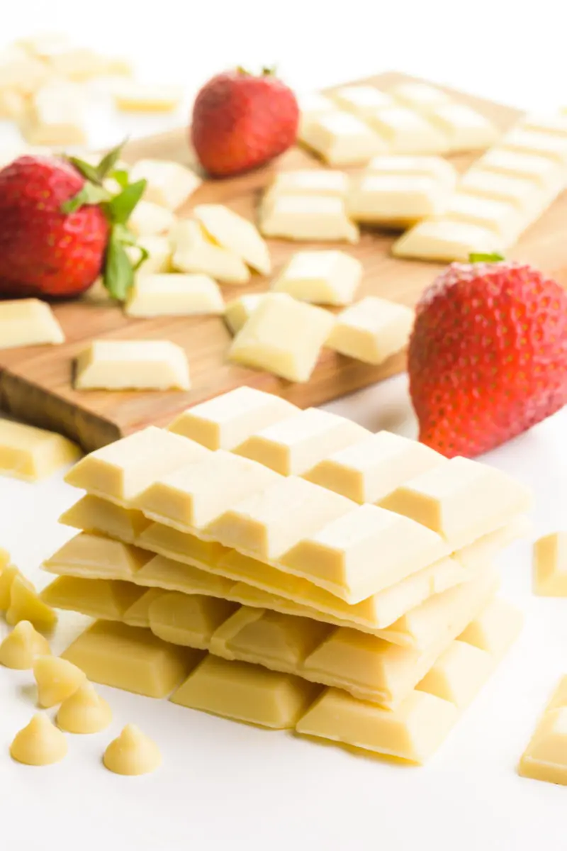 A stack of several vegan white chocolate bars sit in front of a wooden cutting board with more of the chocolate chunks and fresh strawberries.
