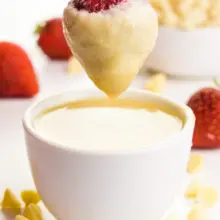 A hand holds a strawberry over a bowl of melted white chocolate. The strawberry is covered in white chocolate. There are white chocolate chips in a bowl behind this and also on the table top. There are fresh strawberries in the background.