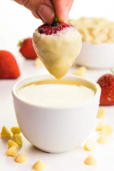 A hand holds a strawberry over a bowl of melted white chocolate. The strawberry is covered in white chocolate. There are white chocolate chips in a bowl behind this and also on the table top. There are fresh strawberries in the background.