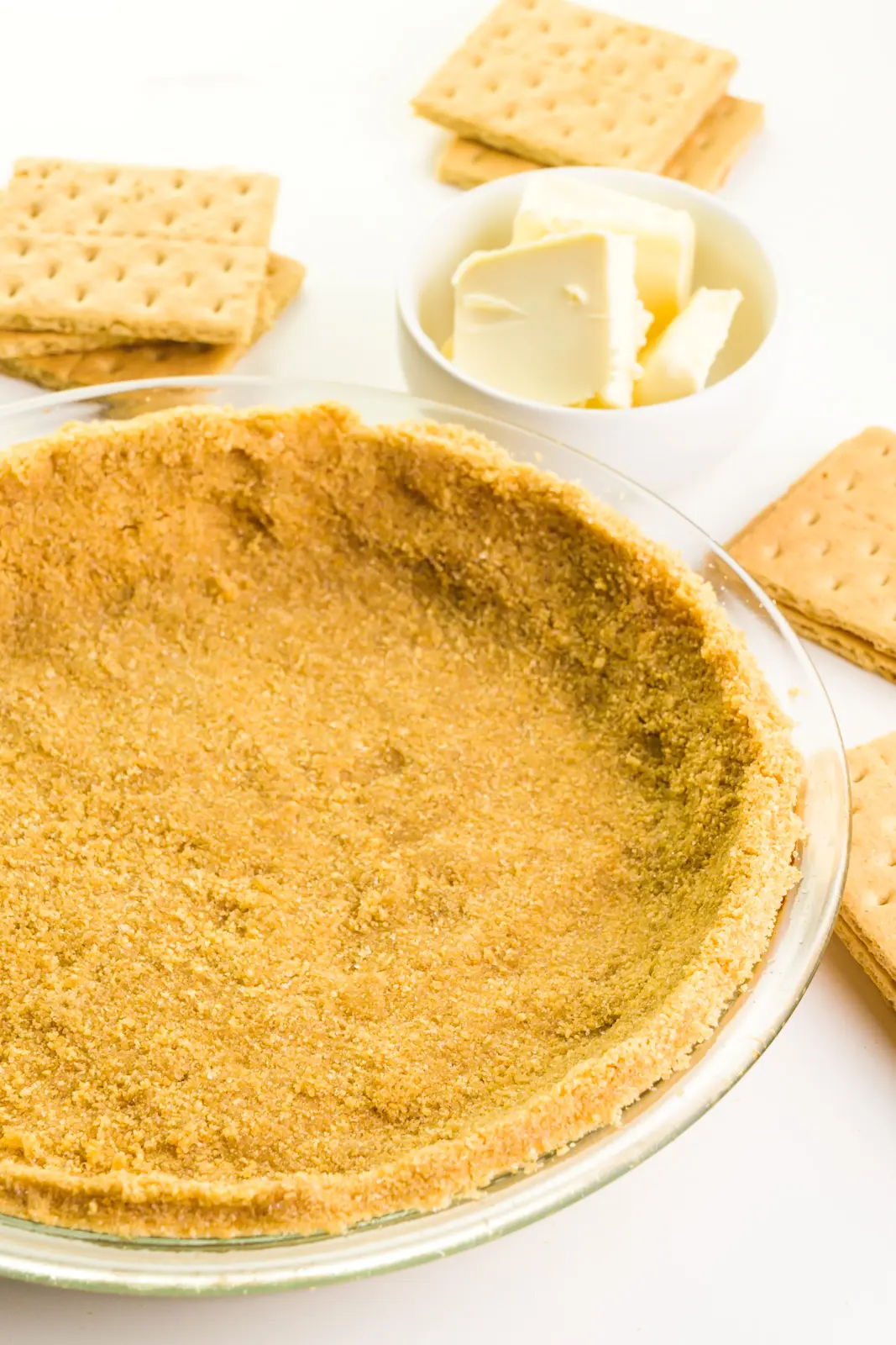 A graham cracker crust is in a glass pie pan. There are graham crackers and vegan butter around it.