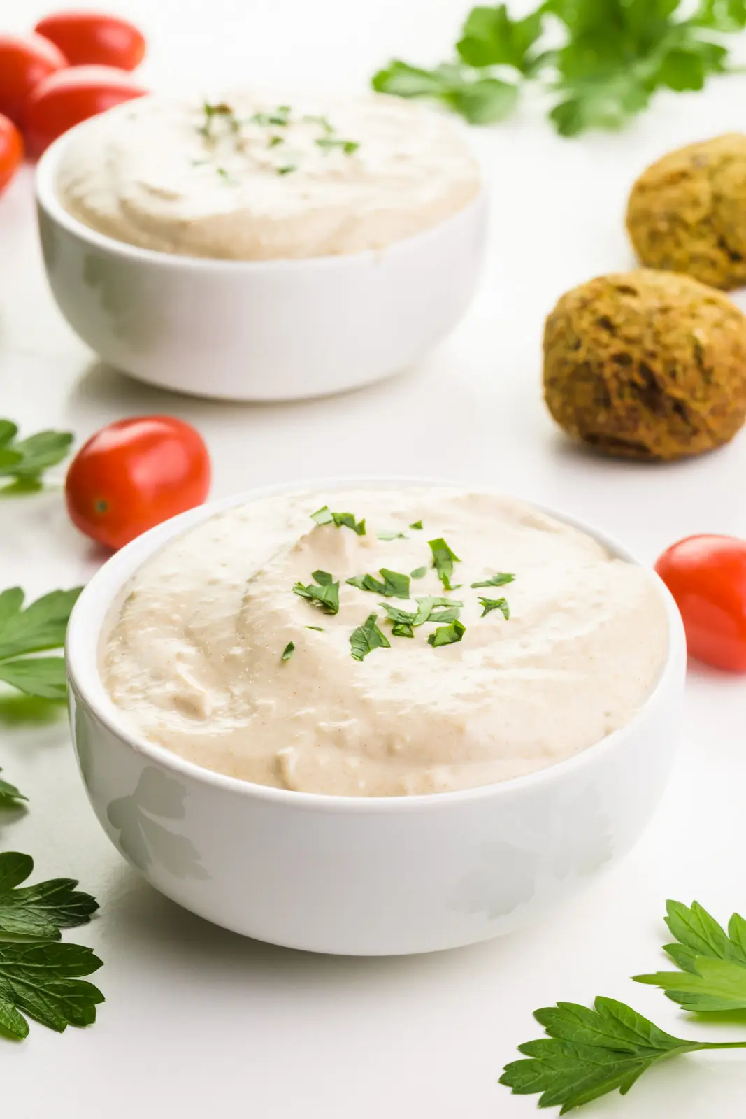 Two bowls of tahini sauce have fresh herbs around them along with red cherry tomatoes and falafel.