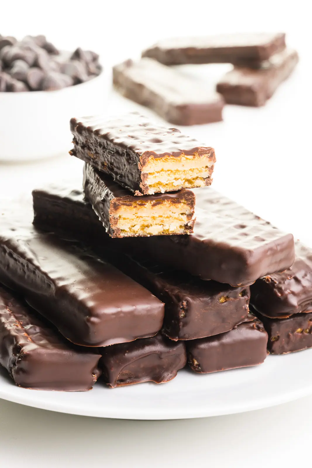 A stack of vegan Kit Kat Bars has two at the top that have been chopped in half, revealing the cookie underneath a chocolate coating. There are chocolate chips and more bars in the background.