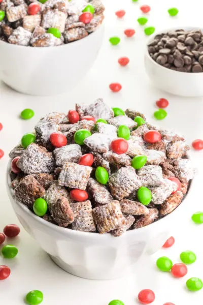 A bowl of Christmas puppy chow has red and green candies around it. There's a bowl of chocolate chips the background and another bowl of the sweet cereal.