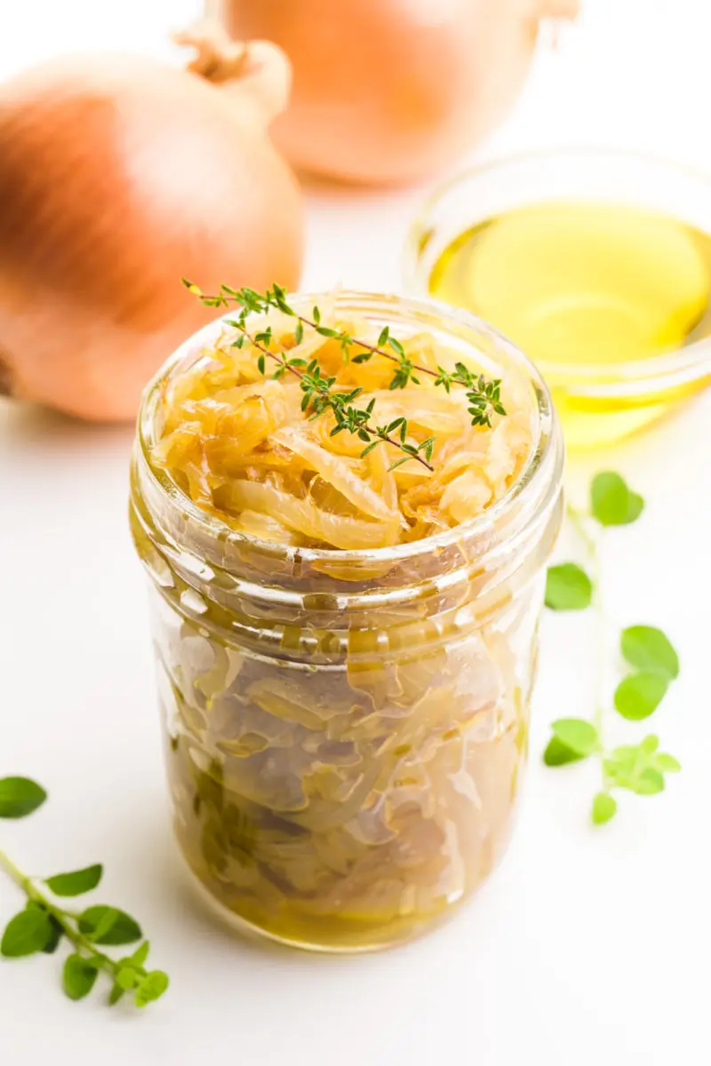 A mason jar holds onion confit with sprigs of thyme on top. There's more herbs around it, and a bowl of olive oil and onions behind it.