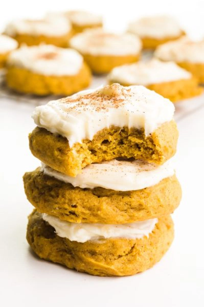 A stack of three vegan pumpkin cookies and the top one has a bite taken out. There are more cookies in the background.