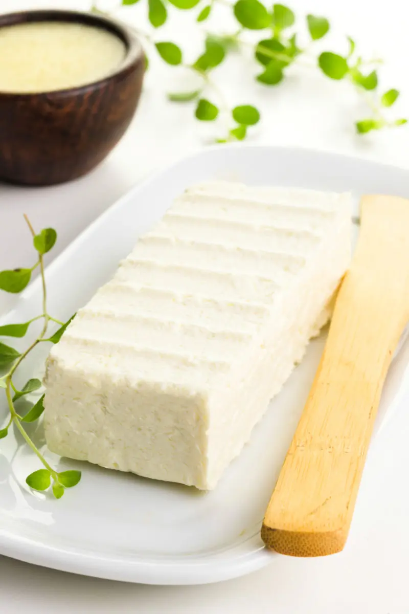 A block of vegan butter sits on a white plate with a wooden butter knife beside it. There are herbs around it with a bowl of melted butter behind it.
