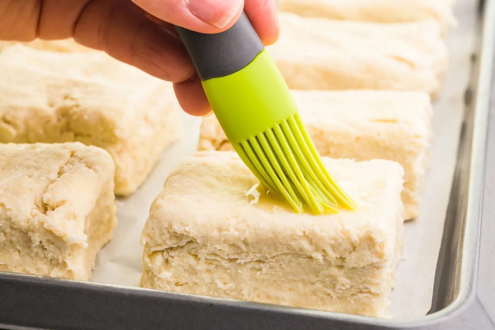 A hand holds a pastry brush, brushing liquid over unbaked biscuits.