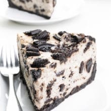 A slice of Oreo cheesecake sits on a plate with a fork beside it. There are two more slices on plates behind it.