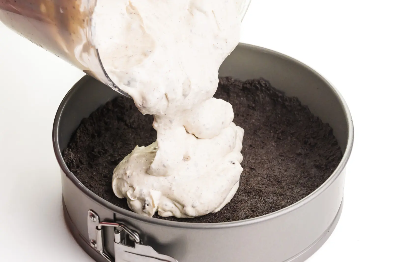 A creamy mixture is being poured into a springform pan with a chocolate cookie crust.
