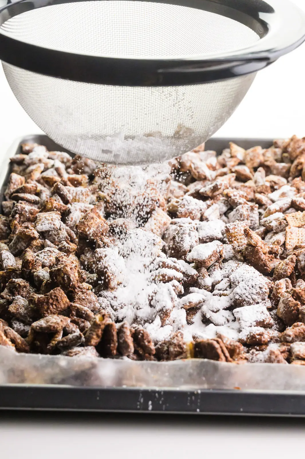 Powdered sugar is being sifted over a pan full of chocolate coated cereal pieces.
