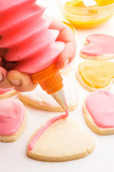 A hand holds a frosting bottle and is outlining a sugar cookie in pink frosting. there are more cookies and frosting behind it.