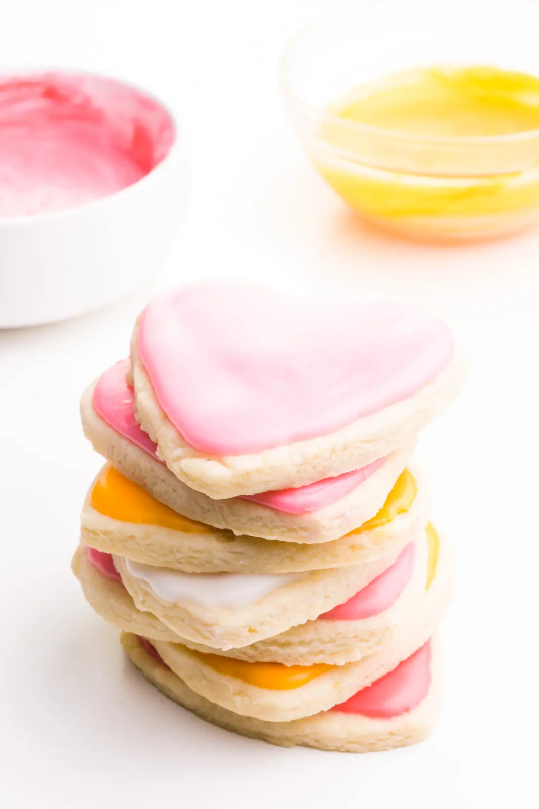 A stack of heart-shaped sugar cookies are frosting in pink and orange. There are bowls of colored frosting behind it.