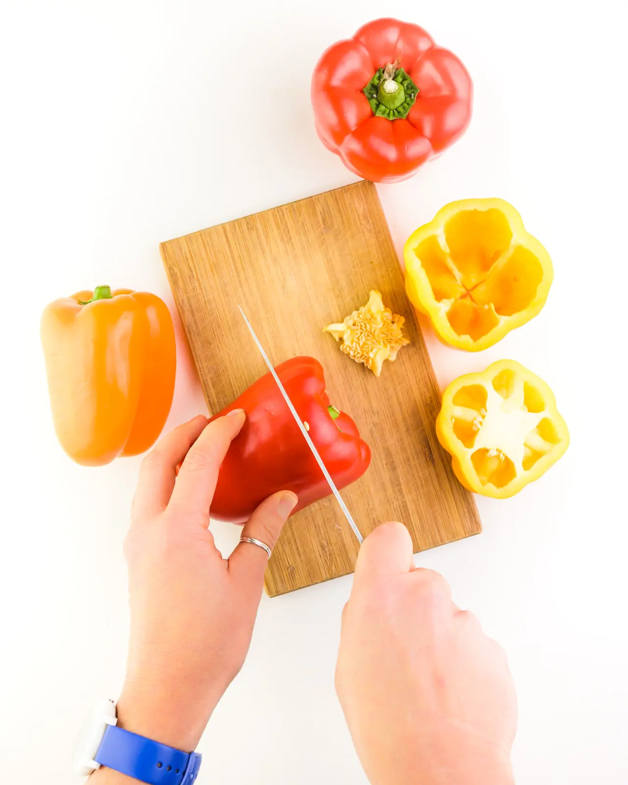 Colorful bell peppers sit on and near a cutting board. A hand holds one pepper while the other hand holds a knife, cutting the top off of it.