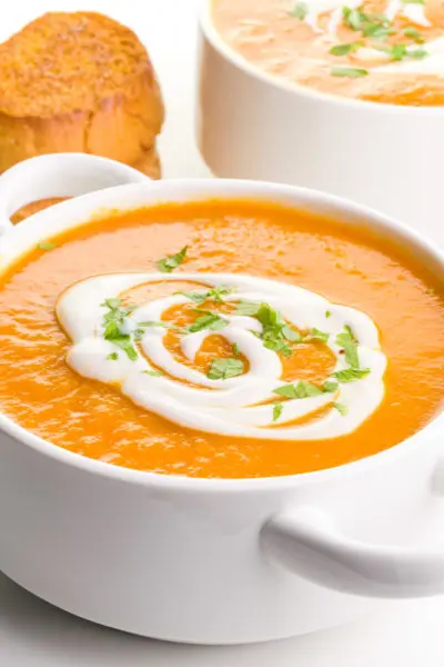 A bowl of carrot ginger soup has cream drizzled over the top and chopped parsley. There's a another bowl of soup behind it along with a stack of toasted bread.