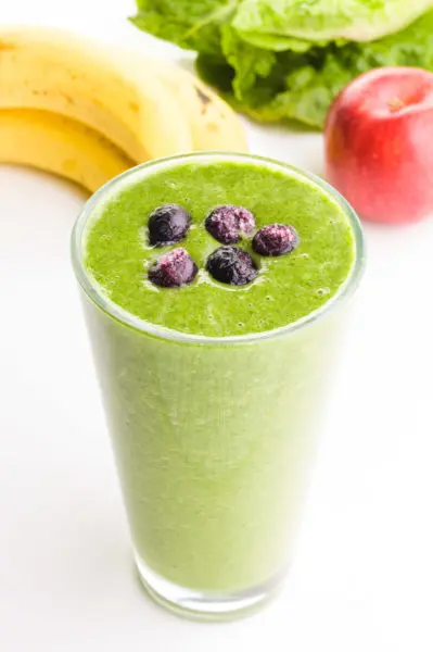 A green smoothie has blueberries on top. There are bananas, apples, and greens behind it.