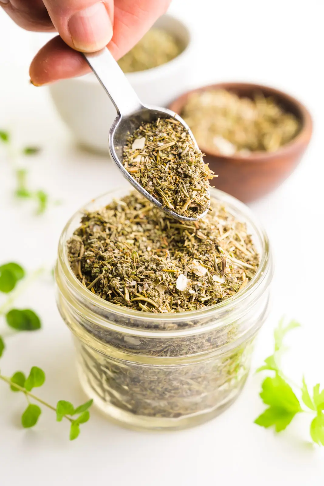A spoon holds seasoning over a bowl of more seasoning. There are fresh herbs and bowls with more seasoning behind it.