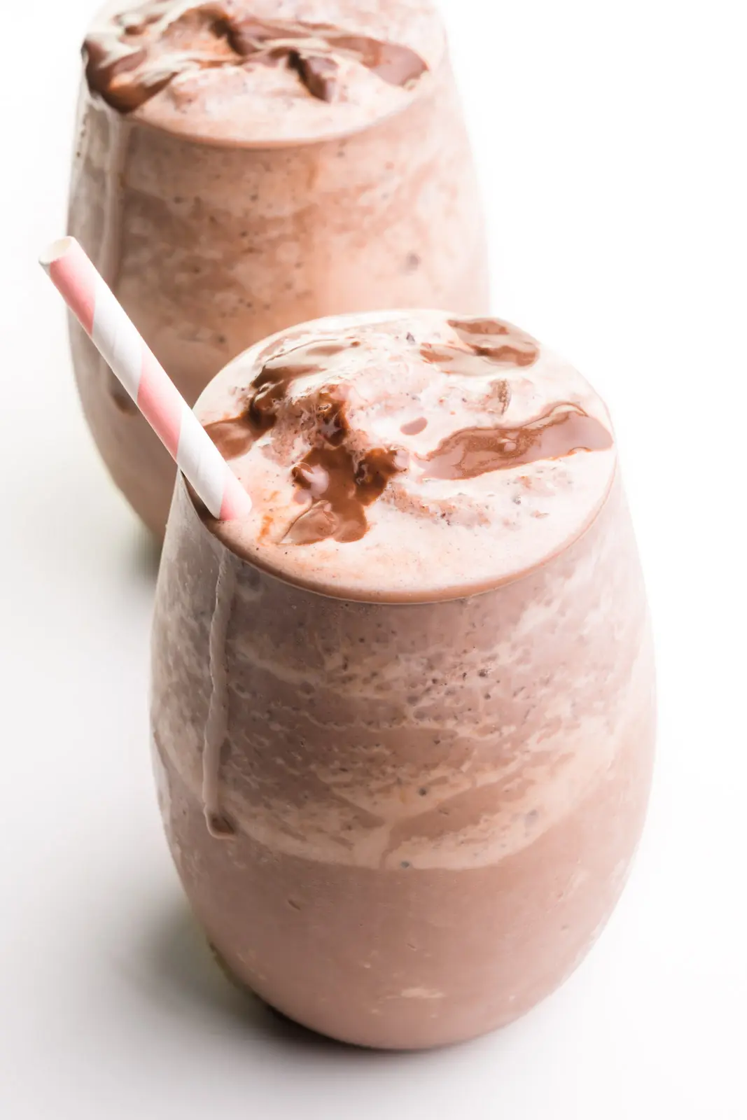 Two glasses hold vegan chocolate milkshakes with chocolate syrup and paper straws.