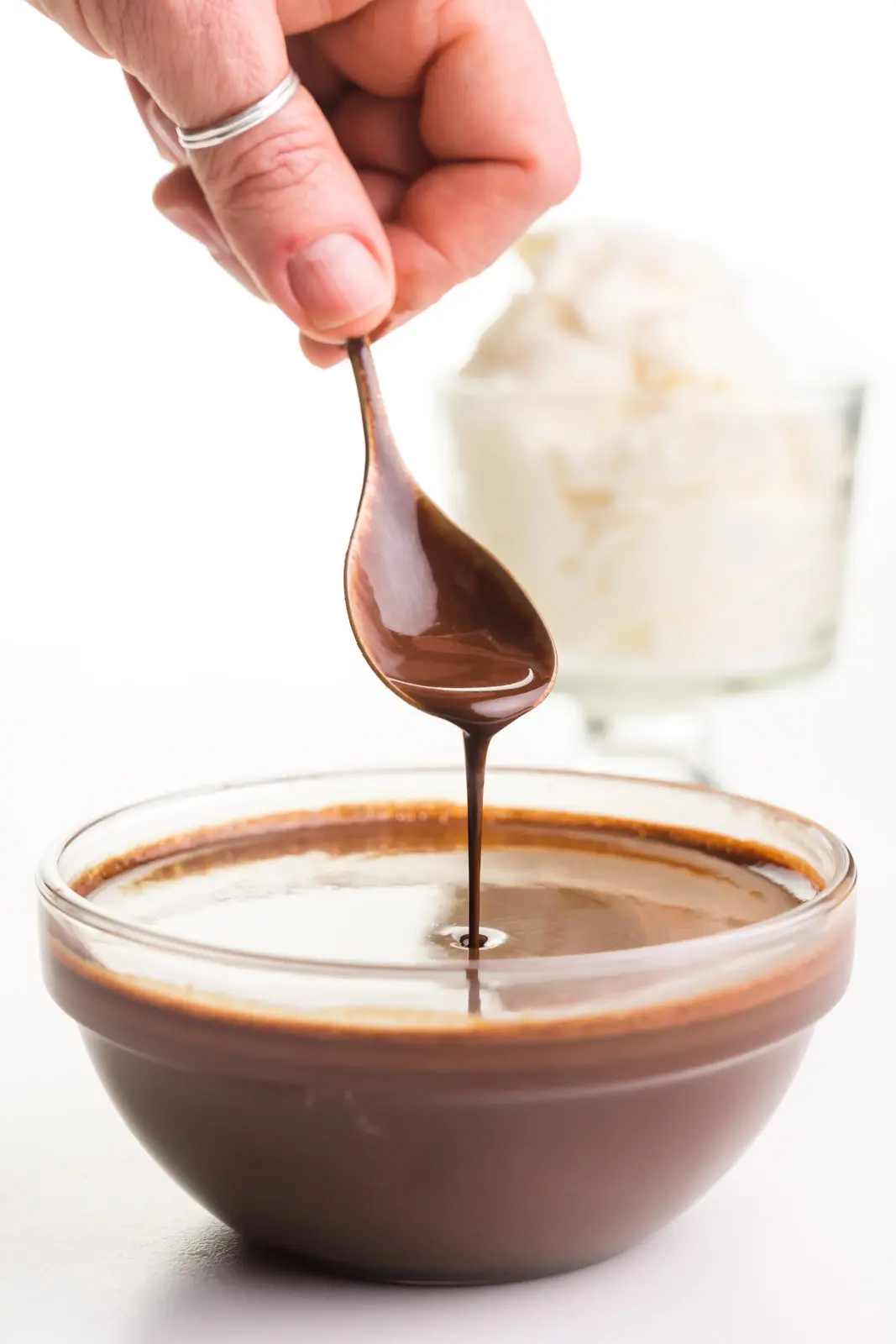 A hand holds a spoon over a bowl of vegan chocolate syrup. There's a bowl of vegan ice cream behind it.