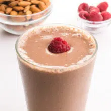 A closeup of a chocolate smoothie in a glass with a fresh raspberry on top. There are bowls of almonds and raspberries in the background.
