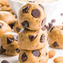A stack of three almond butter balls are studded with chocolate chips. There are more balls in the background, along with almonds, chocolate chips, and a bowl of almond flour.