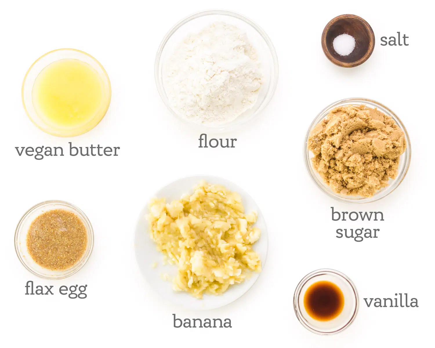 Ingredients are displayed on a table. The labels next to them read, salt, brown sugar, vanilla, banana, flax egg, vegan butter, and flour.