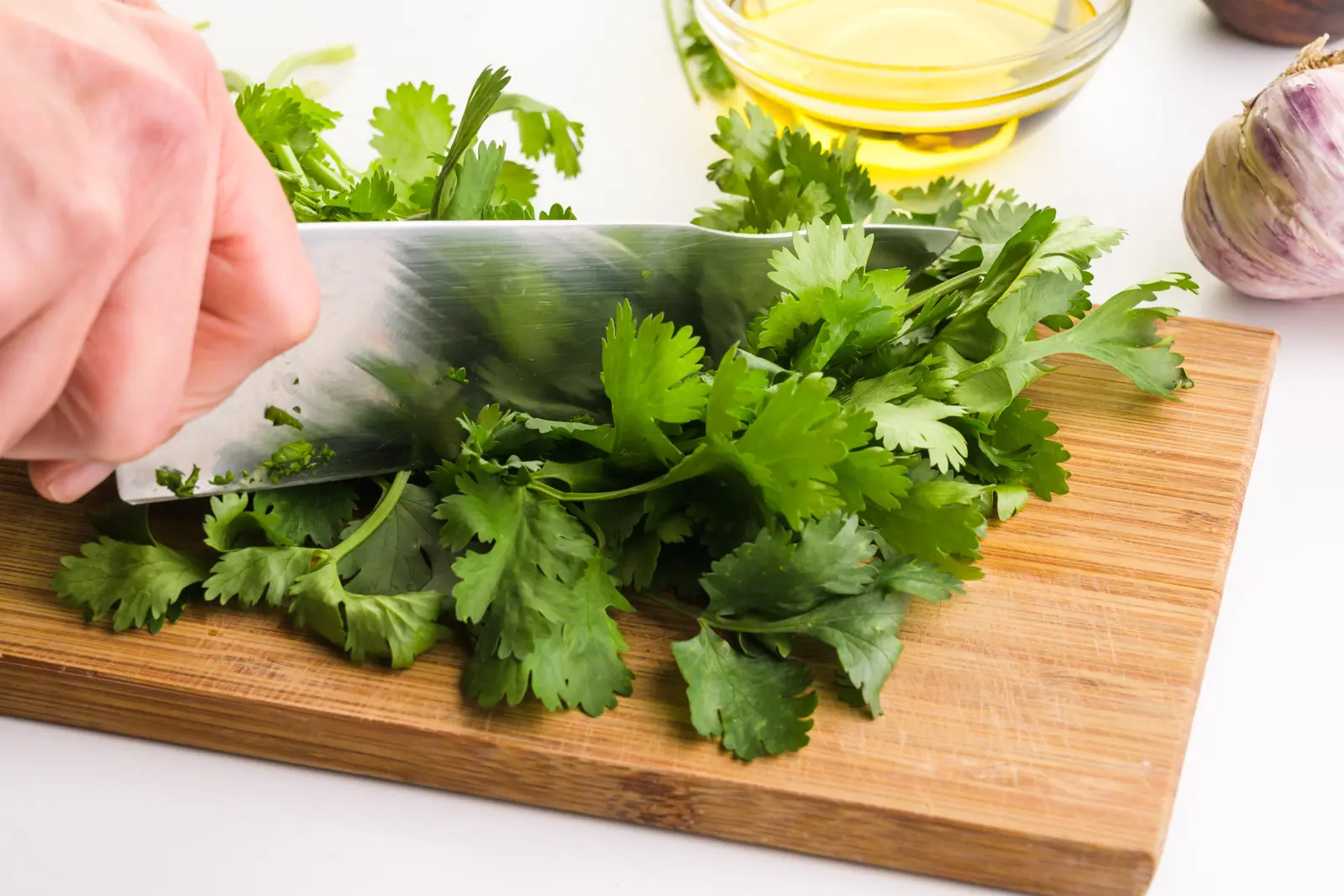 A hand holds a knife cutting cilantro on a wooden cutting board. There other ingredients in the background, such as a bowl of olive oil and garlic.
