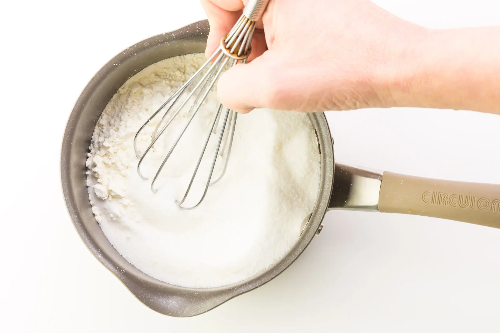 A hand holds a whisk, stirring flour and sugar together in a saucepan.