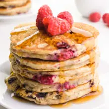 A stack of fruity pancakes with fresh raspberries on top. There is syrup drizzling over the side. There are more pancakes in the background and fresh raspberries.