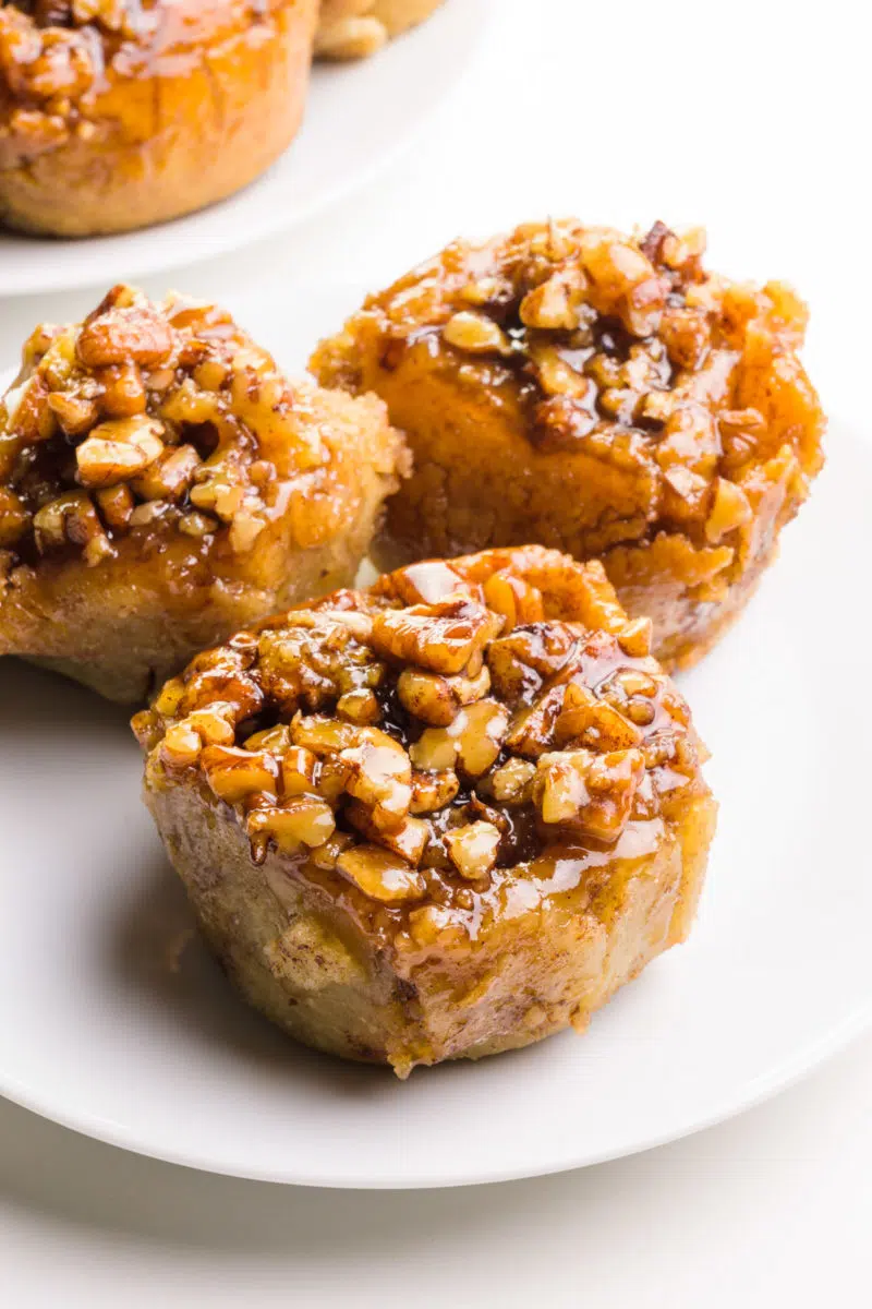 Three gluten-free sticky buns are on a plate. There's another plate of these rolls barely visible in the background.
