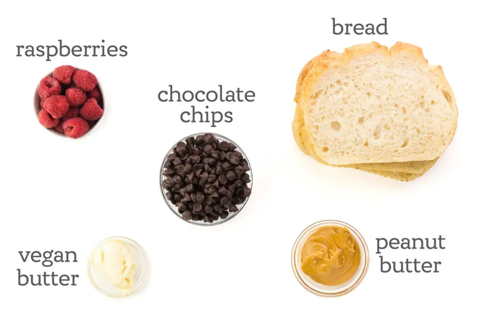 Ingredients are on a white table top. The labels read, "raspberries, chocolate chips, bread, peanut butter, and vegan butter.