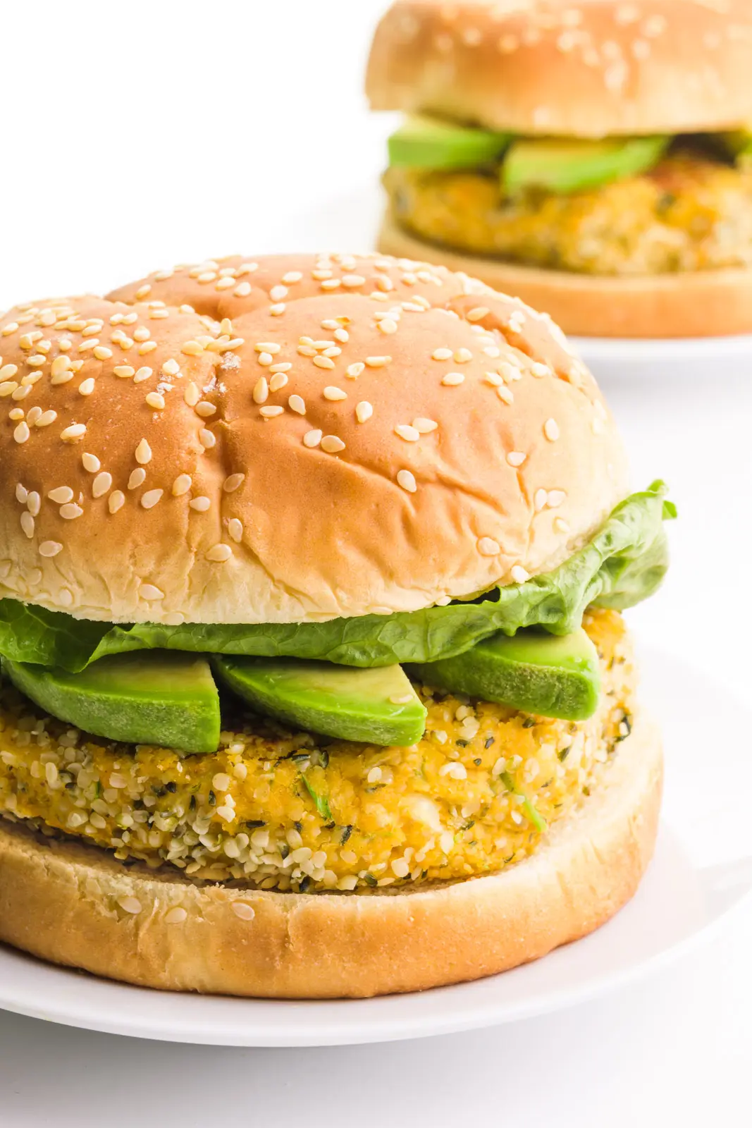A hemp burger sits on a plate. It has avocado slices and lettuce on it. There's another one on a plate behind it.