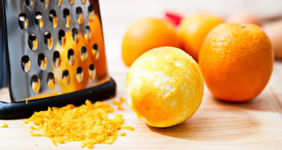 A zested orange sits next to a box grater, orange zest, and more oranges all on a cutting board.