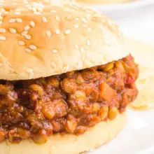 A bun holds lentil sloppy joes on a plate next to potato chips. There's another plate with the same sandwich in the background.