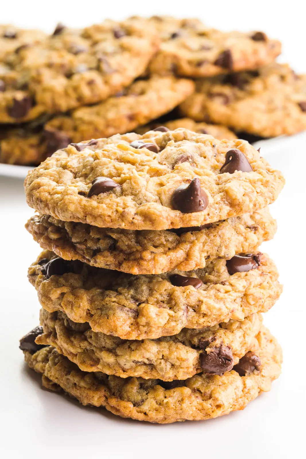 A stack of vegan oatmeal chocolate chip cookies sits in front of a plate with more cookies.