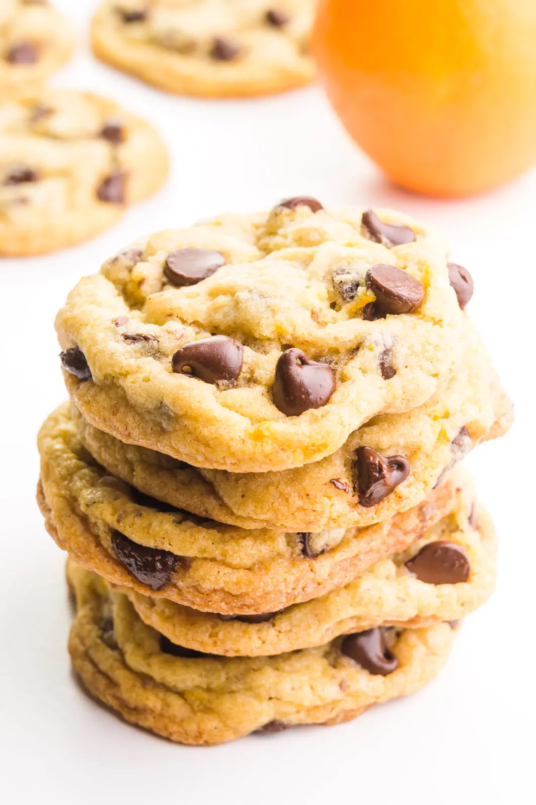 A stack of orange chocolate chip cookies has an orange and more cookies in the background.