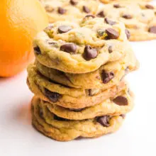 Orange chocolate chip cookies are stacked in front of an orange and more cookies in he background.