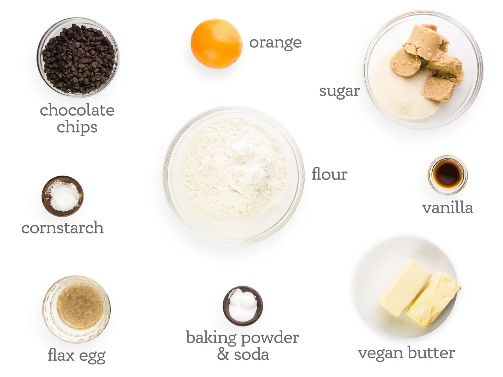 Ingredients are on a white table. The labels read, "orange, sugar, vanilla, vegan butter, baking powder + soda, flax egg, cornstarch, chocolate chips."