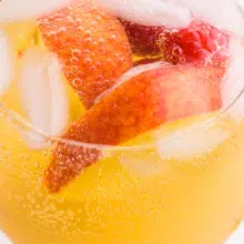 Looking into a wine glass full of peach sangria, ice cubes, raspberries, and peach slices.